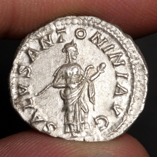 An ancient silver coin held between fingers, featuring a standing figure and raised Latin text around the edge.