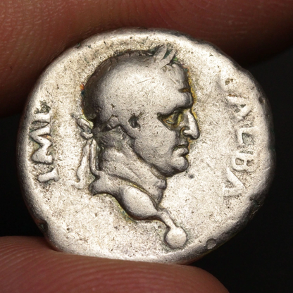An ancient silver coin featuring a profile portrait of a Roman figure, held between two fingers against a dark background.